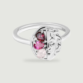 Three red and pink stones sit beside a sculpted face on a polished silver ring against a light grey background.