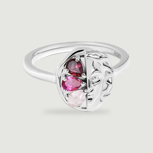 Three red and pink stones sit beside a sculpted face on a polished silver ring against a light grey background.