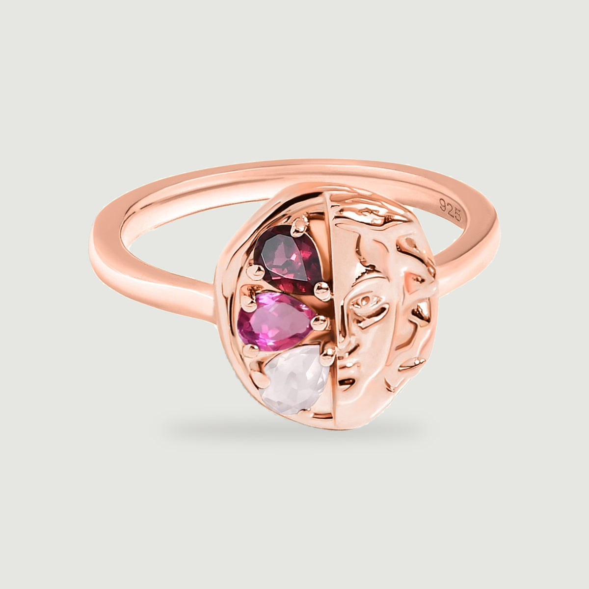 A rose gold ring shows a rounded sculpted face beside dark red, bright pink and pale pink stones.