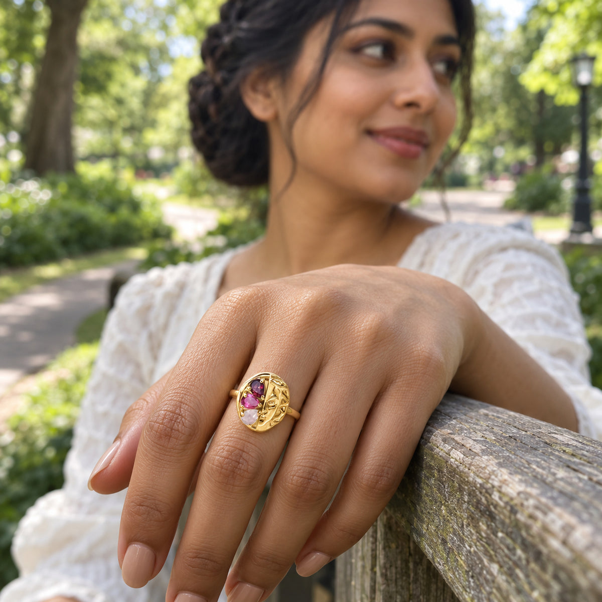 A yellow gold ring with three red and pink pear-shaped stones is worn on a hand resting on a wooden railing in a leafy park.