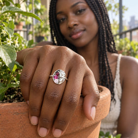 A silver ring with a sculpted face and three colored stones is worn on a hand resting on a terracotta planter beside green leaves.