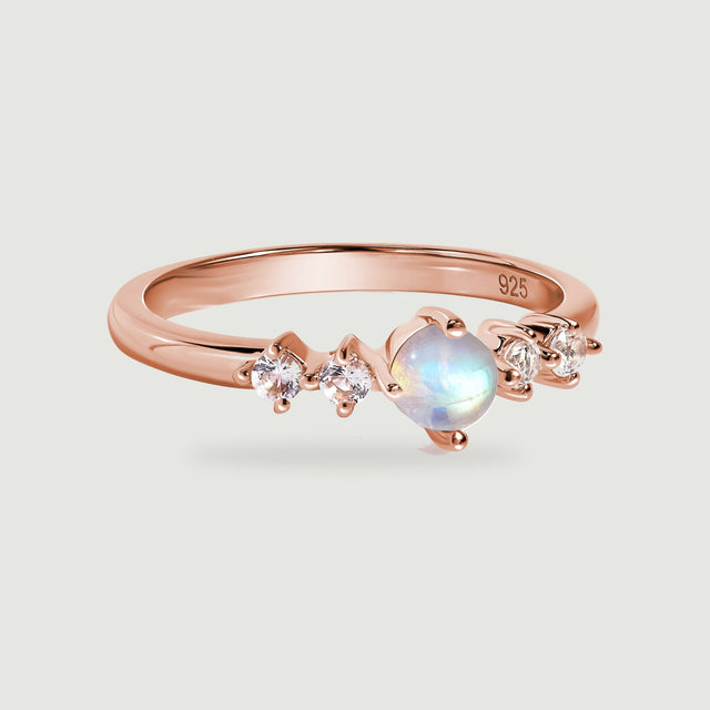 A rose gold ring with a round moonstone and four clear side stones rests on a light gray surface.