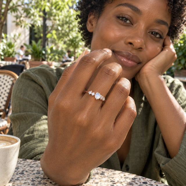 A woman with short curls wears the rose gold moonstone ring at a leafy outdoor cafe.