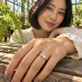 The rose gold moonstone ring is worn on a hand resting on a wooden table among greenhouse plants.