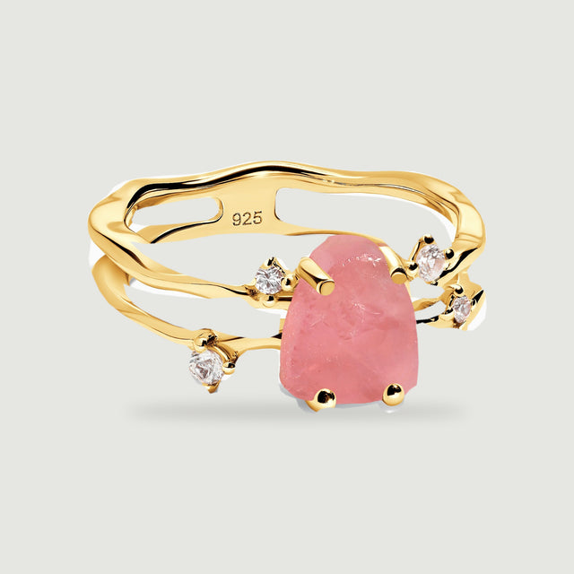 A yellow gold vermeil ring holds a rough pink rose quartz stone beside four clear accent stones on a light gray surface.