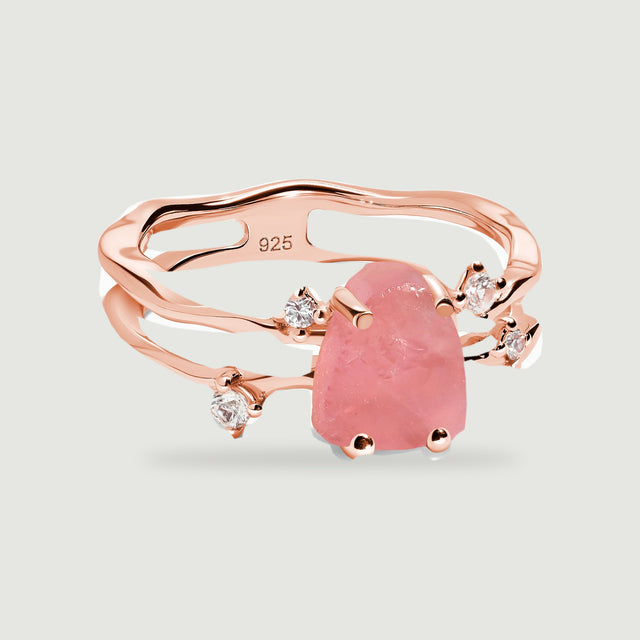 A rose gold vermeil ring with a raw pink center stone and clear round accents rests on a light gray background.