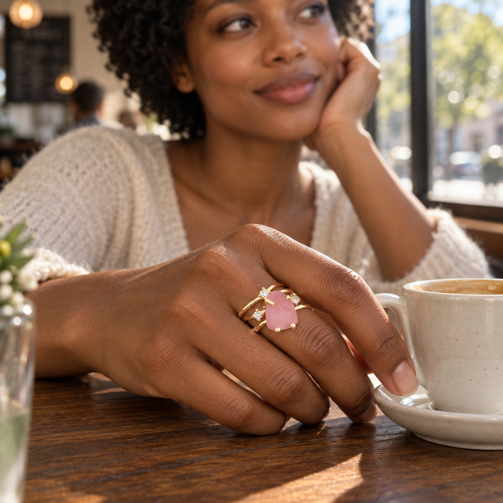 A woman wears a yellow gold vermeil rose quartz ring with clear accents beside a ceramic cup on a wooden cafe table.