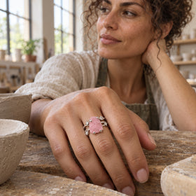 A pink rose quartz ring with a silver split band is worn on a hand resting among ceramic bowls in a pottery studio.