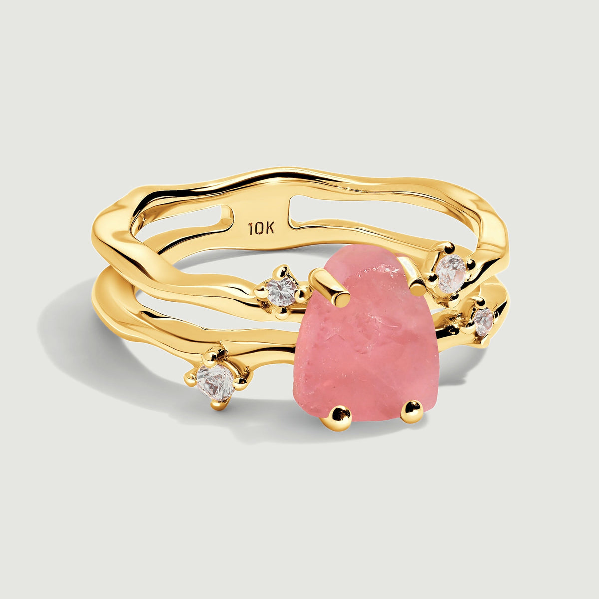 A complete yellow gold ring with a rough pink rose quartz center and four clear accent stones rests on a light gray surface.