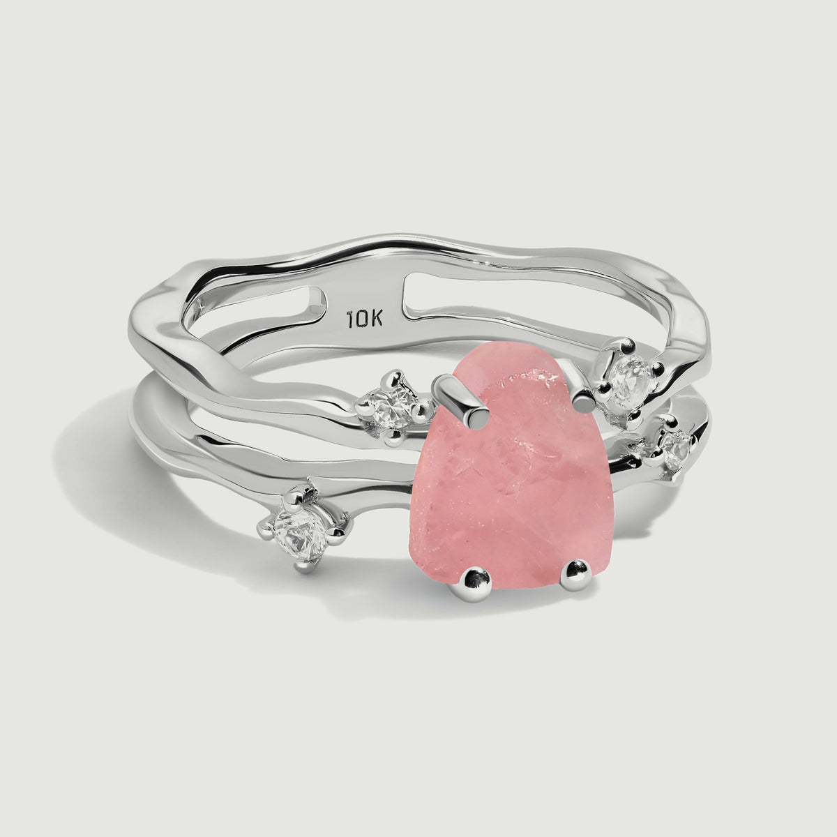 A white gold ring shows its pink rose quartz, rounded prongs, and open double band against a pale gray background.