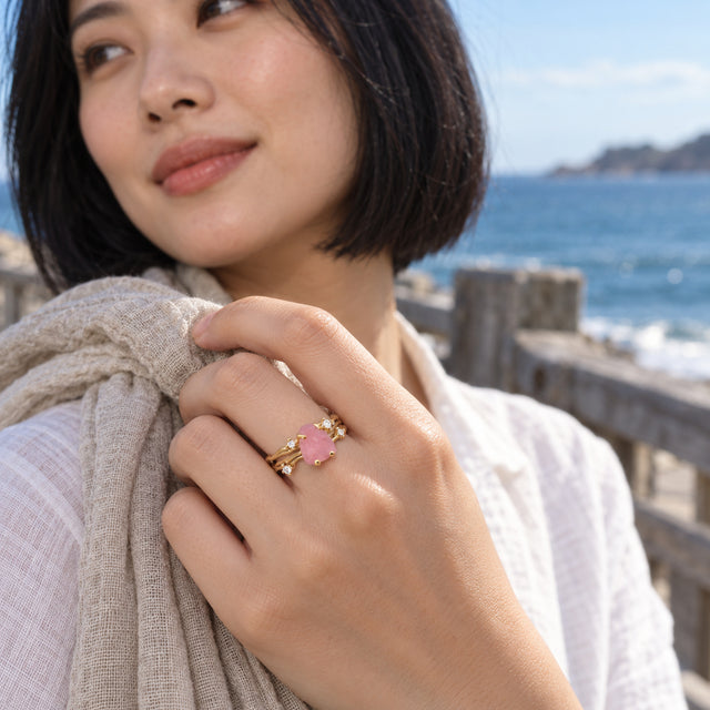 A yellow gold ring with a pink crystal and clear accent stones is shown on a hand holding a linen scarf beside the sea.