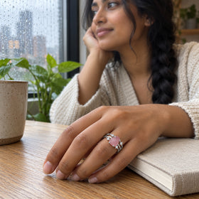A white gold rose quartz ring is visible on a hand resting beside a cloth-covered book near a rain-speckled window.