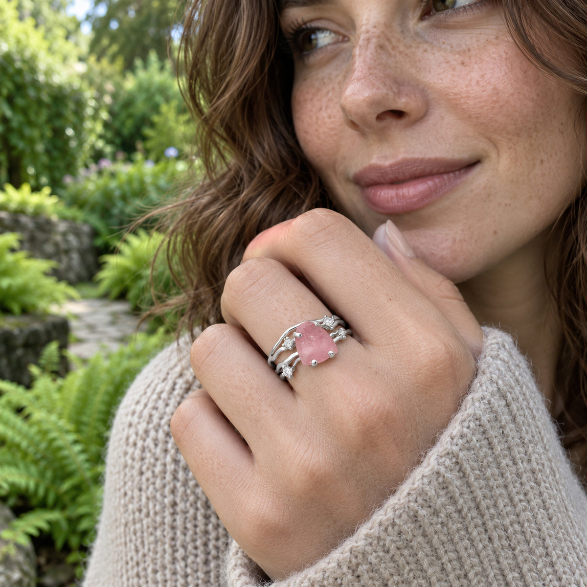 A woman in a knit sweater holds her hand near her chin, showing a white gold pink crystal ring against garden greenery.