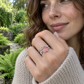 A woman in a knit sweater holds her hand near her chin, showing a white gold pink crystal ring against garden greenery.