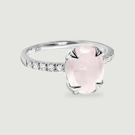 A sterling silver ring with an oval pale pink stone and clear shoulder stones rests on a light gray surface.