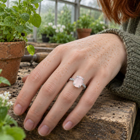 A silver ring with a pale pink oval stone is worn on a freckled hand beside potted plants in a greenhouse.