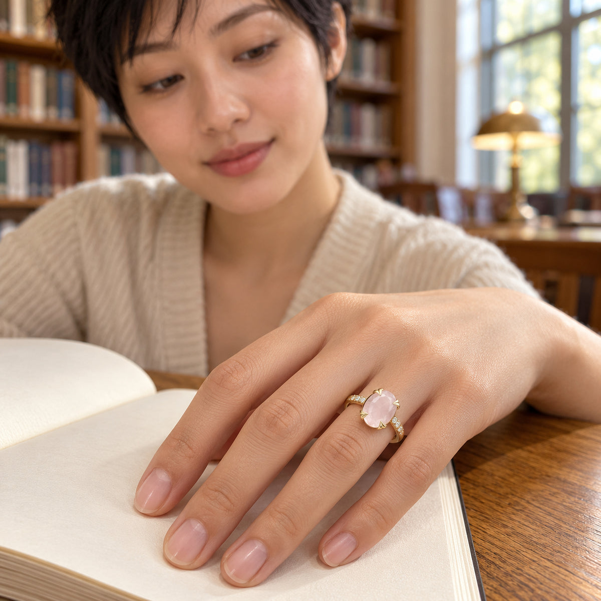A yellow gold vermeil ring is shown on a hand resting across an open book at a wooden library table.