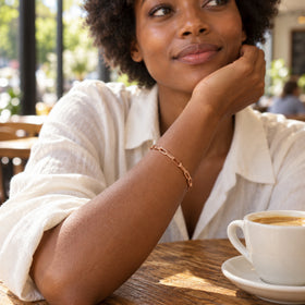 A rose gold chain bracelet is worn on a wrist beside a coffee cup in a sunlit cafe.