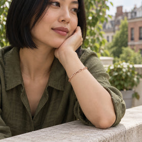 A woman in an olive linen blouse wears a rose gold chain bracelet on a leafy terrace.