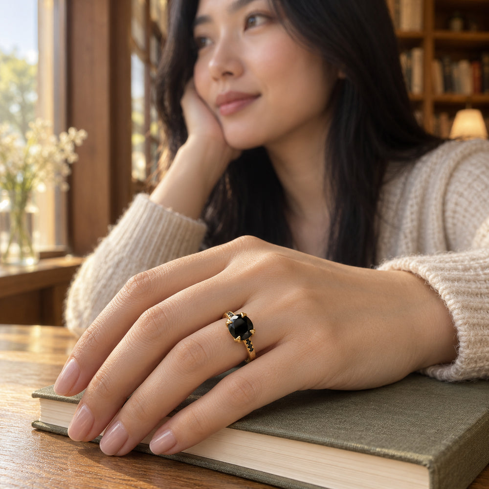 A woman wears the yellow gold ring with its oval black stone while resting her hand on a closed book beside a window.
