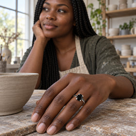 The silver ring sits on a woman's hand beside a clay bowl in a softly lit pottery studio.