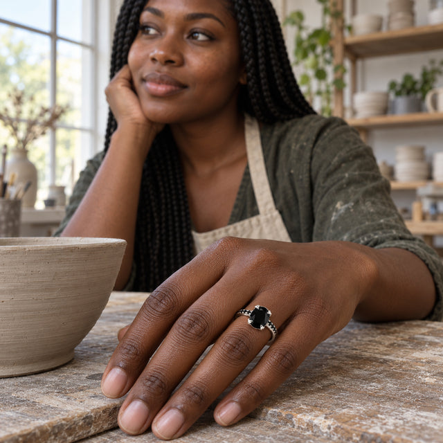 The silver ring sits on a woman's hand beside a clay bowl in a softly lit pottery studio.