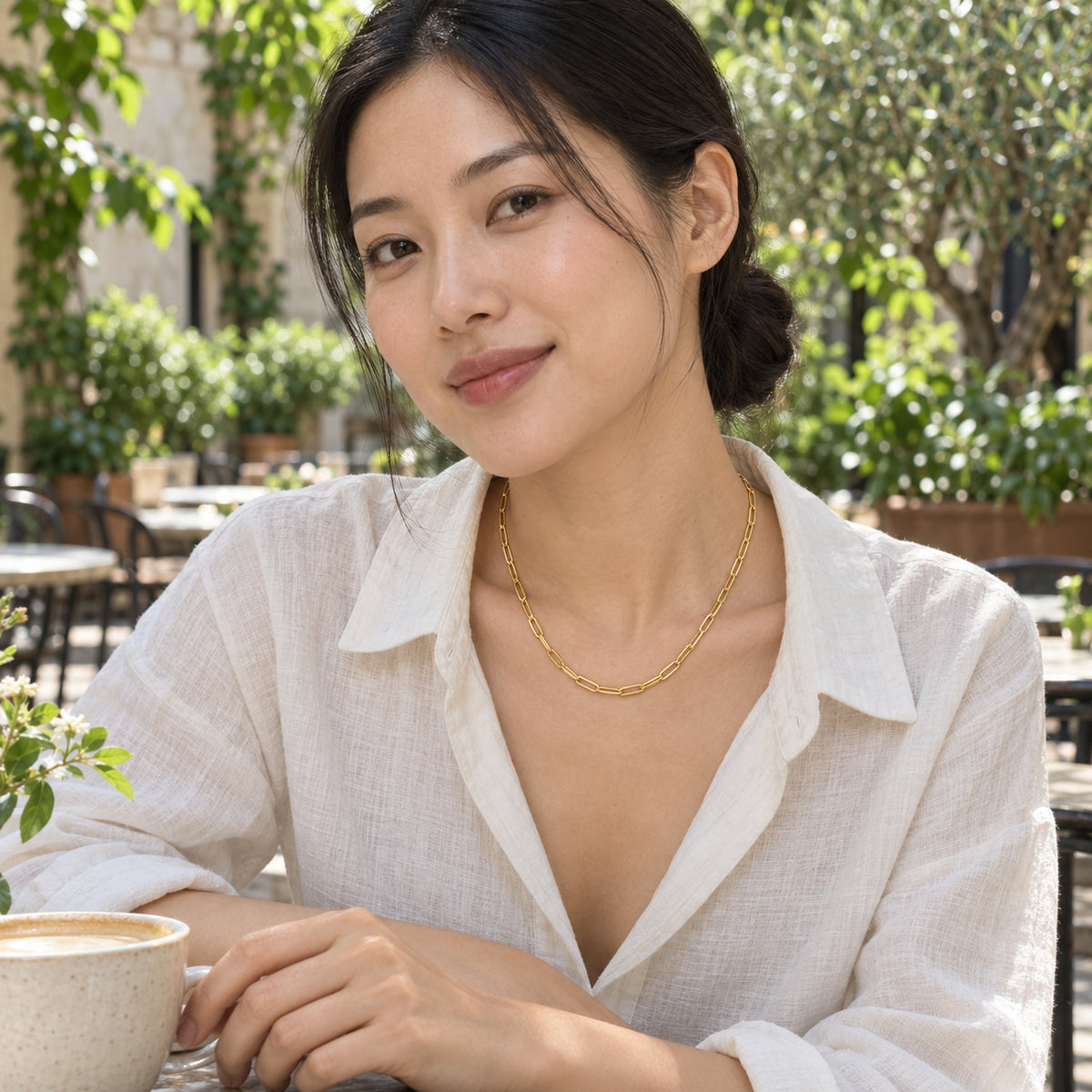 A woman in an ivory linen shirt wears a yellow gold link necklace at a leafy courtyard cafe.