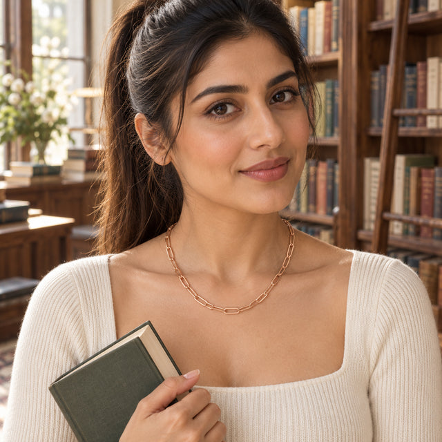 A woman holding a green book wears a rose gold chain necklace above a cream square neckline.