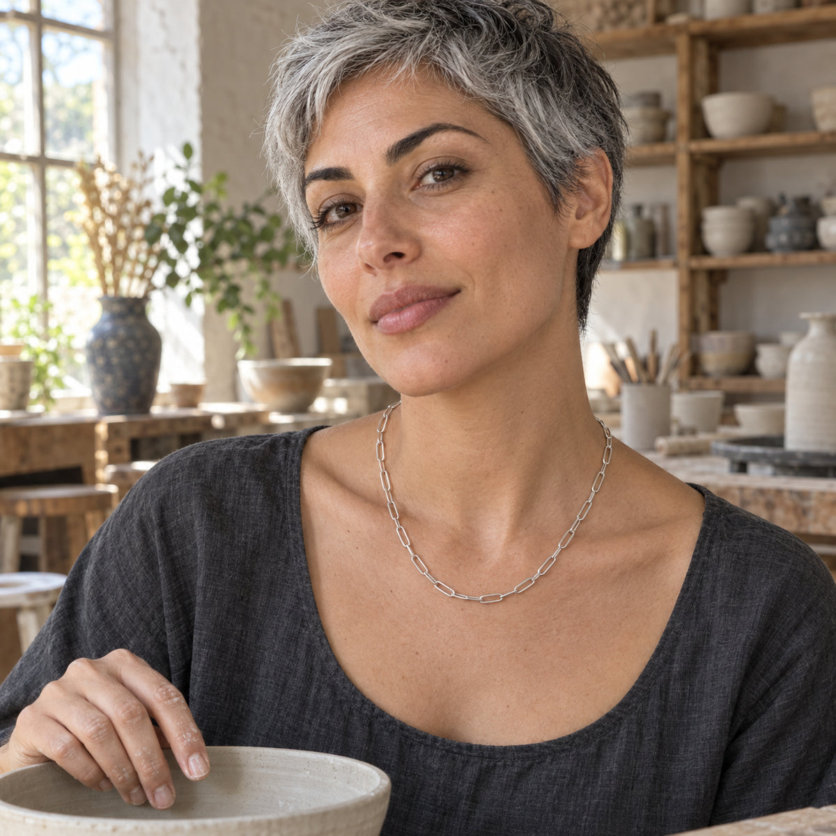 A woman with short gray hair wears a silver link necklace while holding a ceramic bowl in a pottery workshop.