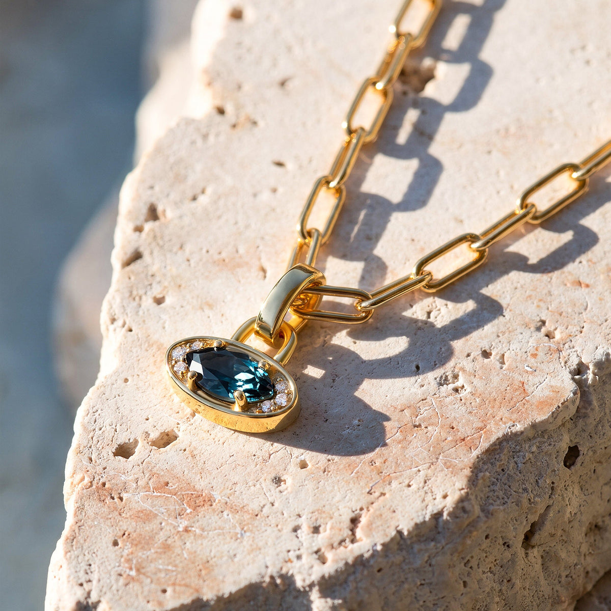 A yellow gold-tone necklace lies on textured pale stone with the blue pendant and link shadows visible.