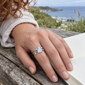 A silver moonstone ring is worn on a freckled hand beside an open sketchbook on a wooden bench.