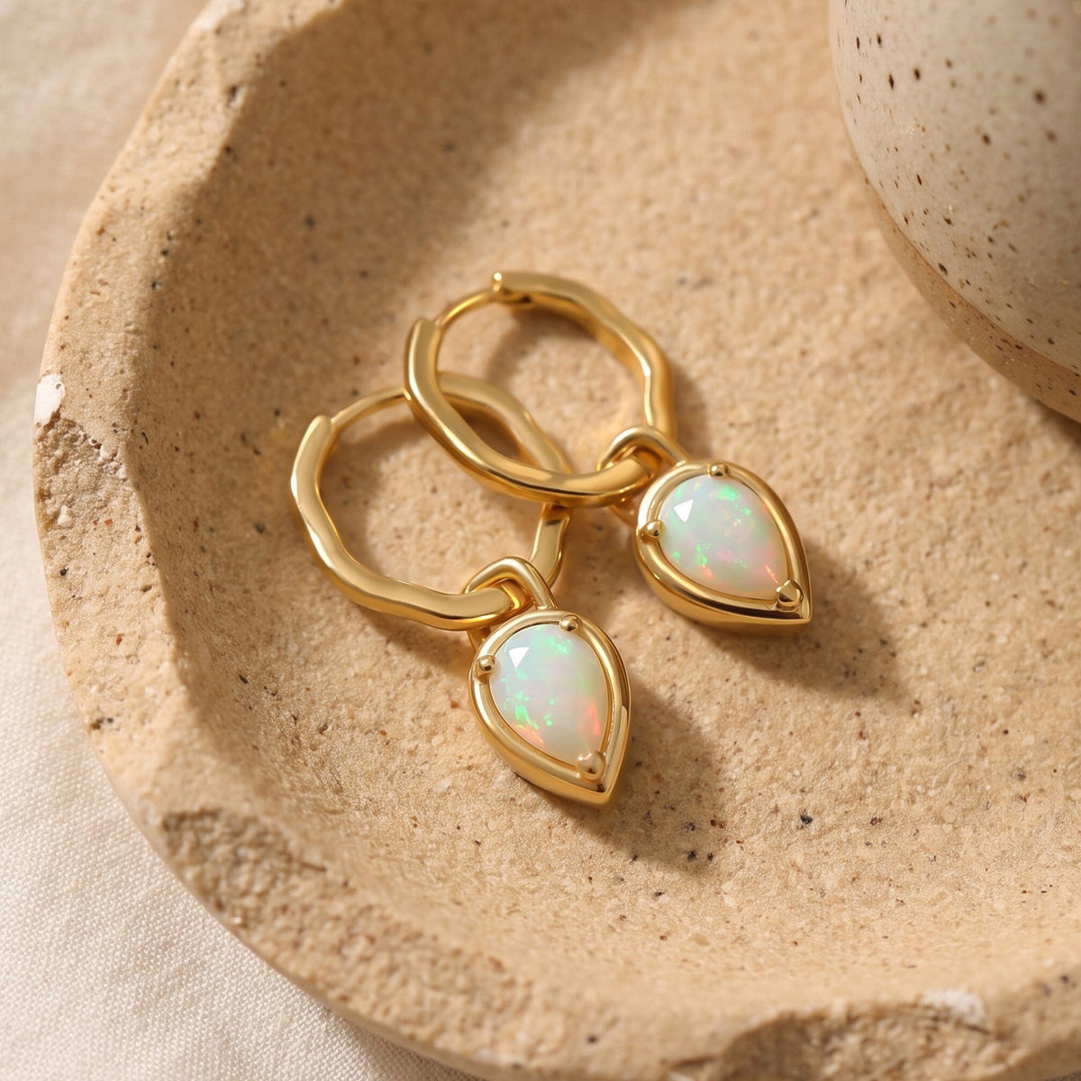 Yellow gold opal hoop earrings lie together on a textured beige ceramic tray.