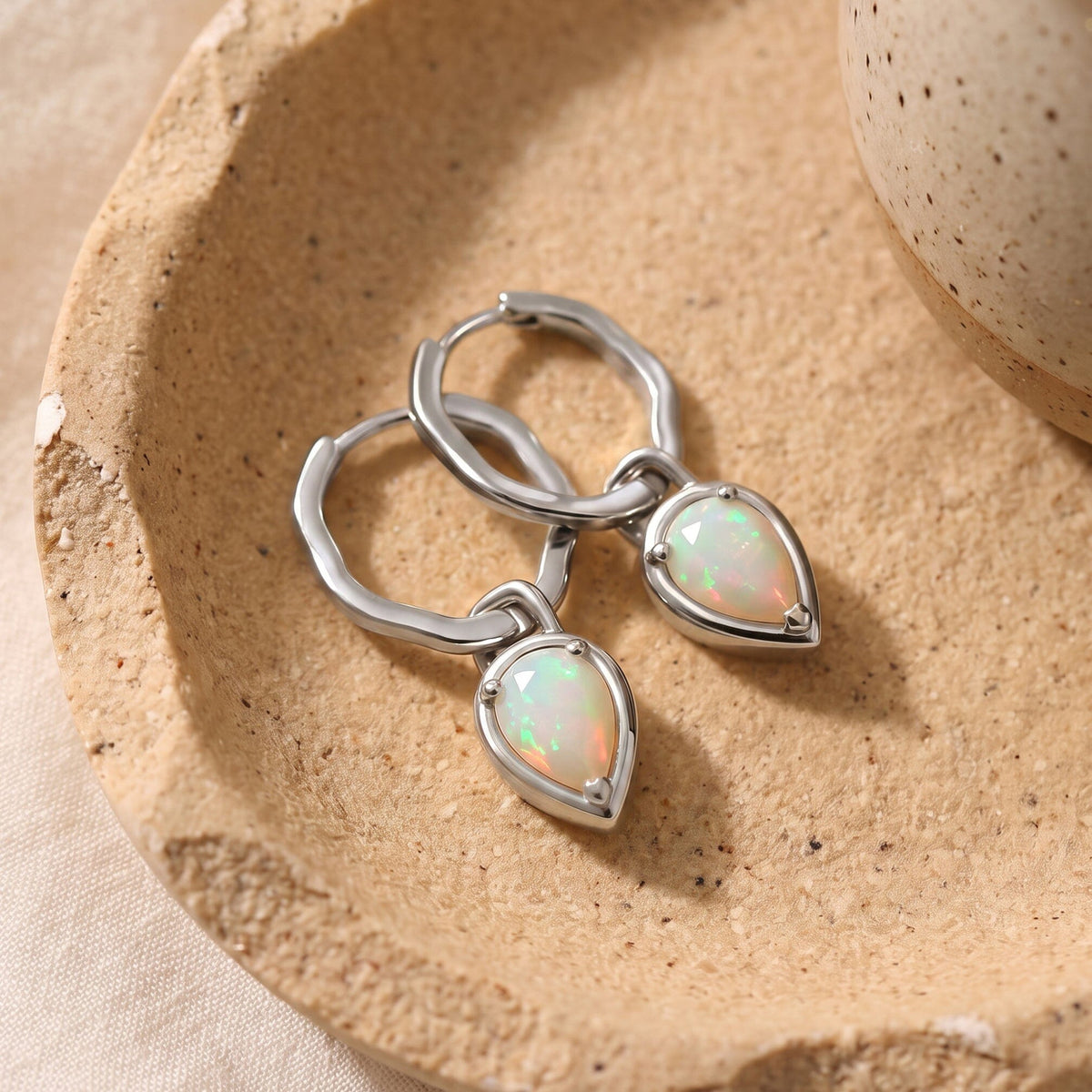 Silver-tone hoops and milky opal drops lie on a speckled stone-colored dish beside a ceramic vessel.