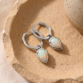 Silver-tone hoops and milky opal drops lie on a speckled stone-colored dish beside a ceramic vessel.
