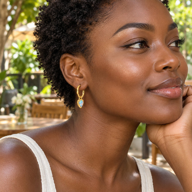 A woman with short natural curls wears a yellow gold opal hoop at a leafy outdoor cafe.