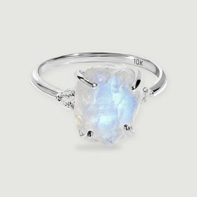 A white gold ring with a rough blue-white moonstone and two round side stones rests on a light gray surface.