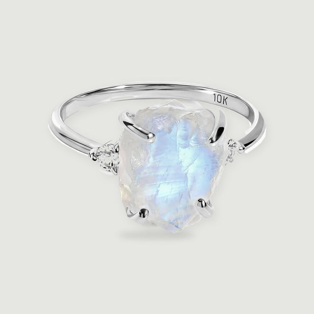 A white gold ring with a rough blue-white moonstone and two round side stones rests on a light gray surface.