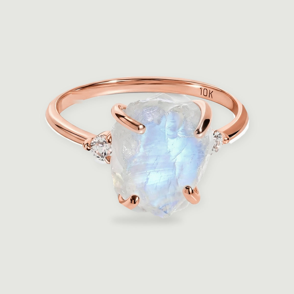 A rose gold ring holds an irregular pale moonstone between rounded prongs and clear side stones.