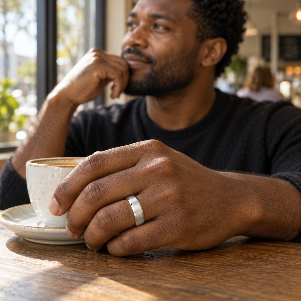A man wears a brushed silver band on his ring finger beside a ceramic coffee cup in a sunlit cafe.
