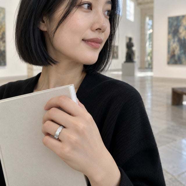 A woman with a black bob wears a silver band while holding a plain cloth-covered book in an art gallery.