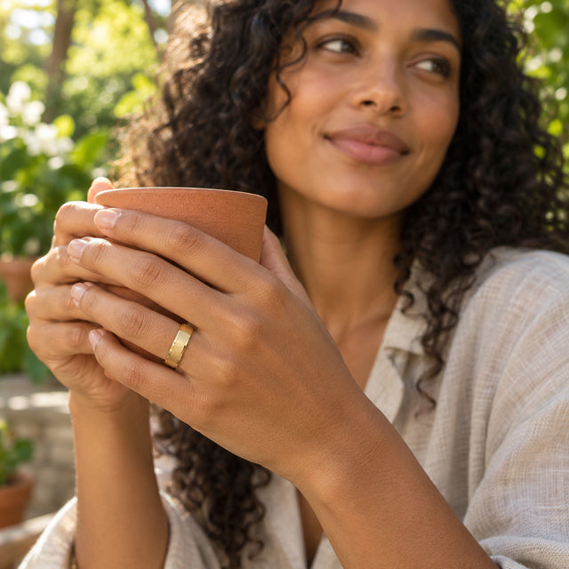 A curly-haired woman wears a yellow gold band as she holds a terracotta cup among green garden leaves.