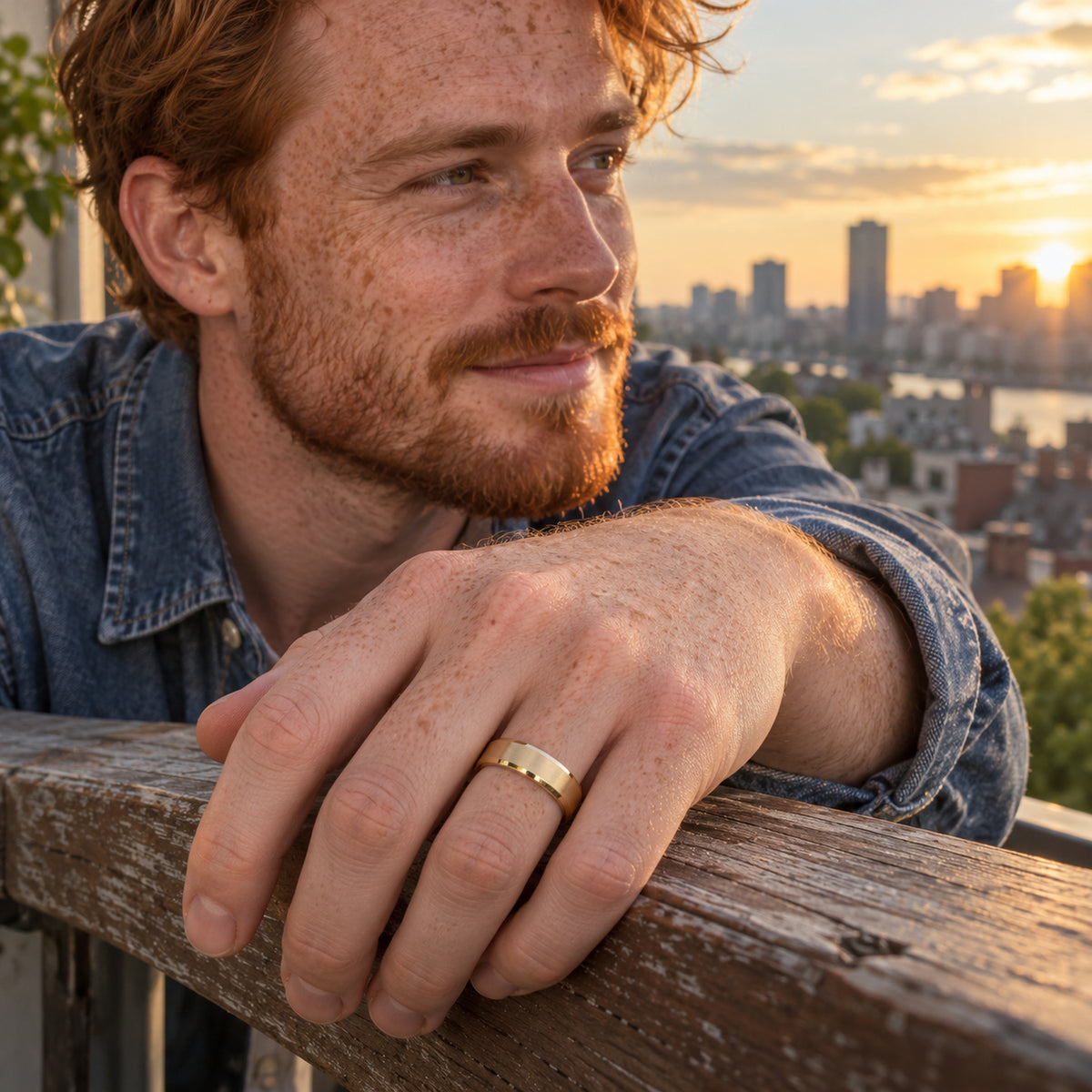 A red-haired man wears a brushed yellow gold ring while resting his hand on a wooden balcony railing at sunset.
