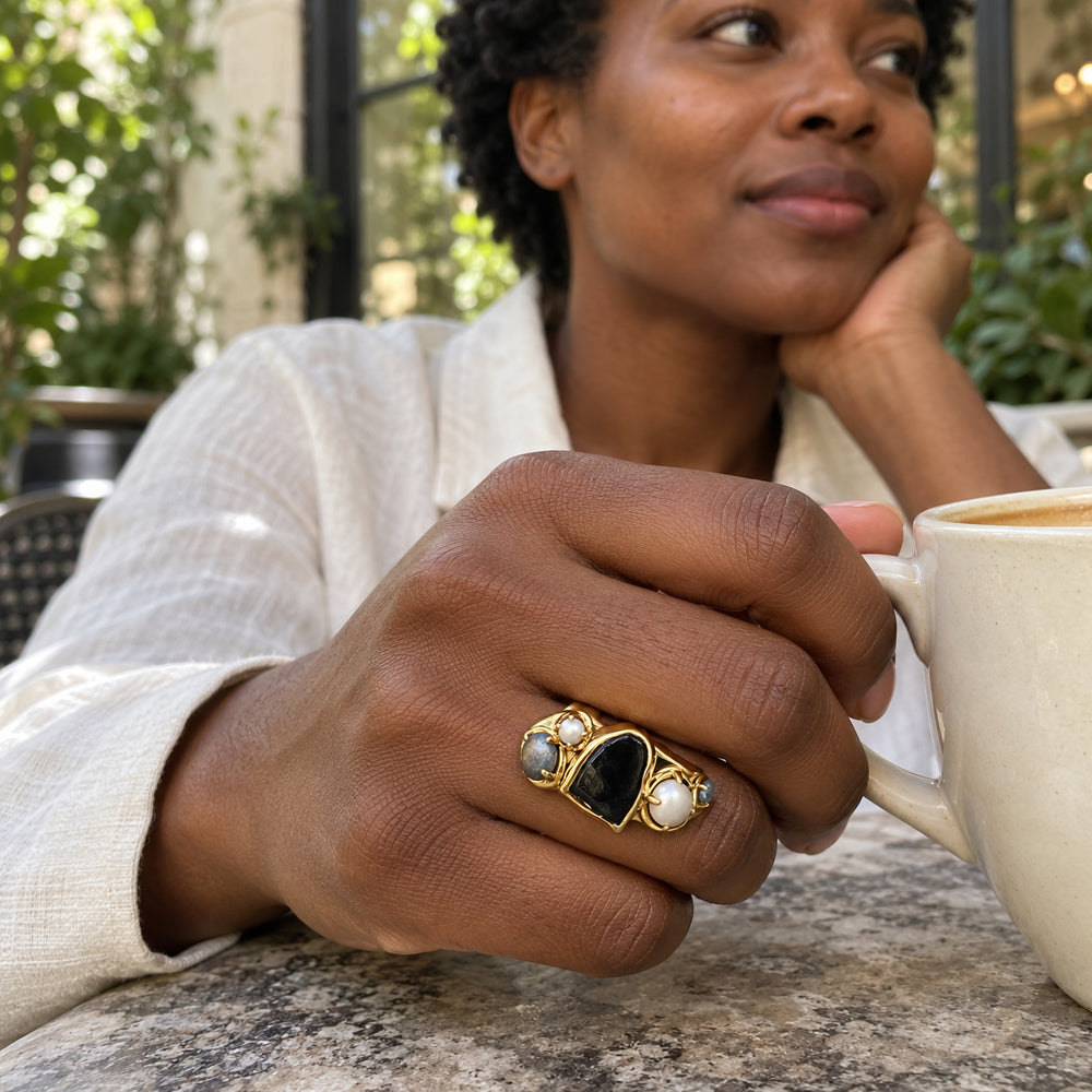 A woman in an ivory linen shirt wears the gold ring while holding a ceramic cup at a leafy courtyard cafe.
