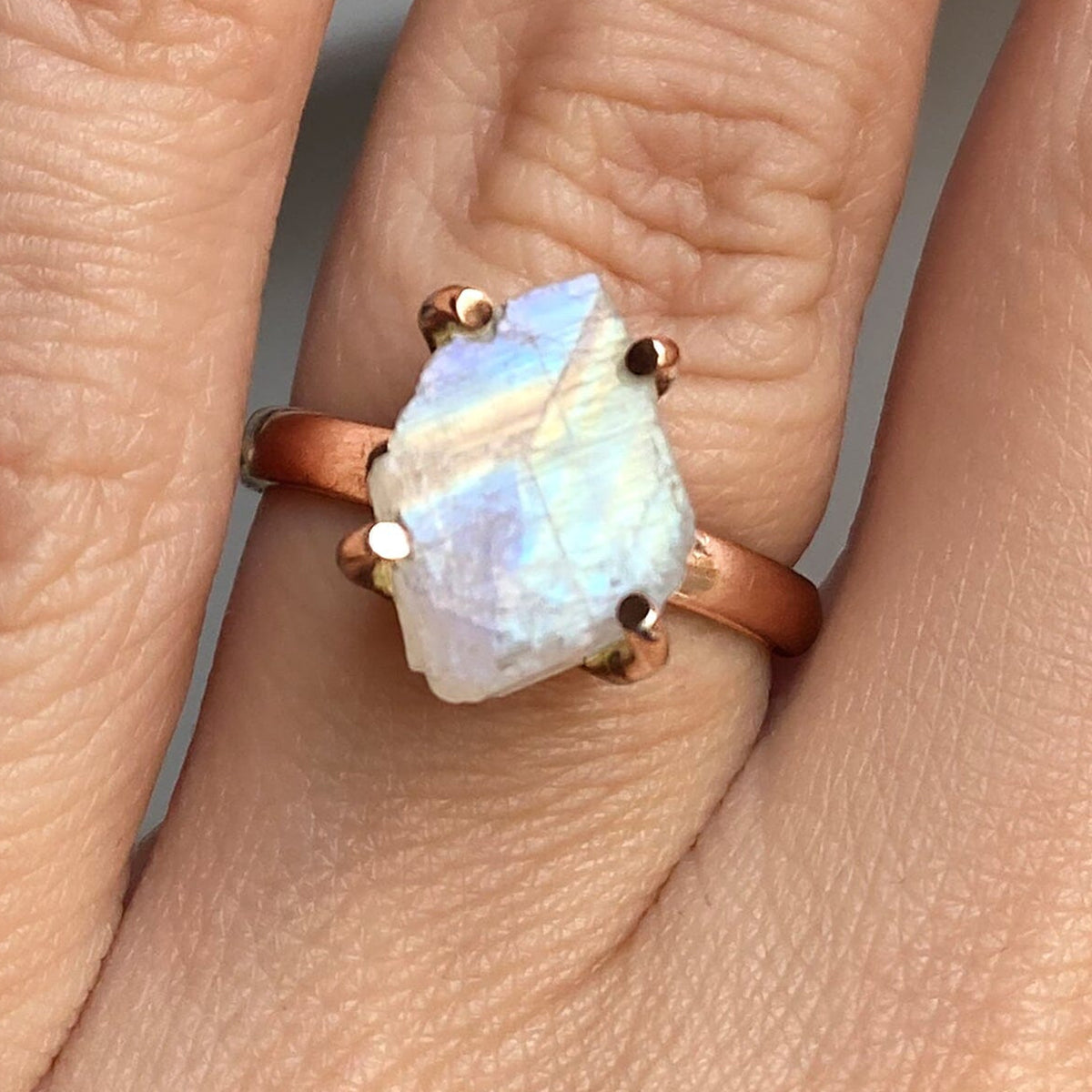 Blue and pale yellow reflections cross a rough moonstone in a rose gold ring shown close up on a finger.