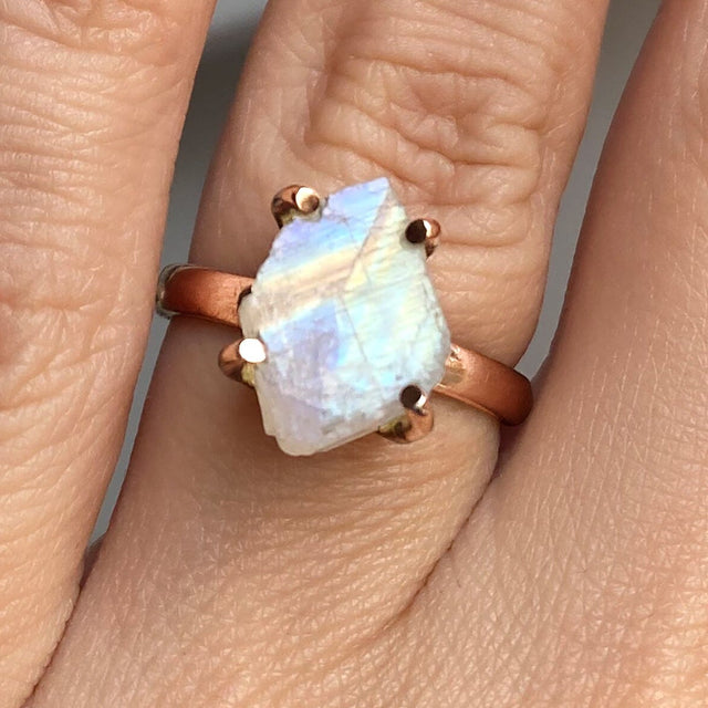 Blue and pale yellow reflections cross a rough moonstone in a rose gold ring shown close up on a finger.