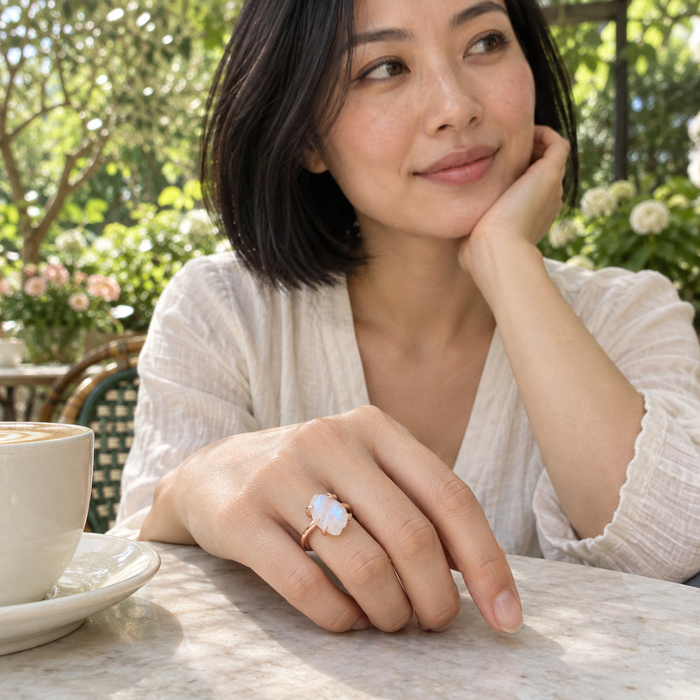 A woman with a black bob wears the rose gold moonstone ring beside a ceramic cup at a leafy garden cafe.