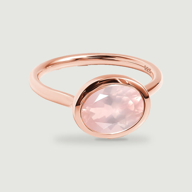 A rose gold ring with a pale pink oval faceted stone rests on a light gray surface.