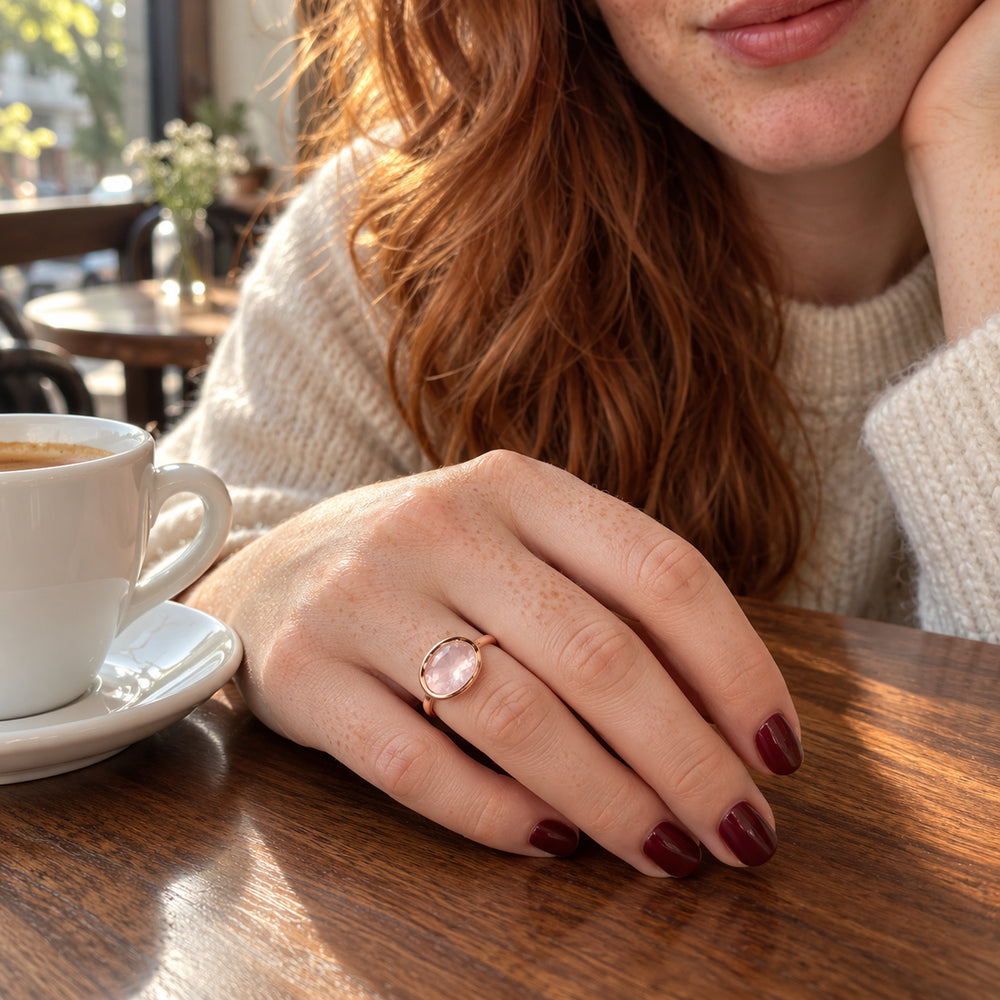 A rose gold ring with an oval pink stone is worn on a freckled hand beside a white coffee cup.