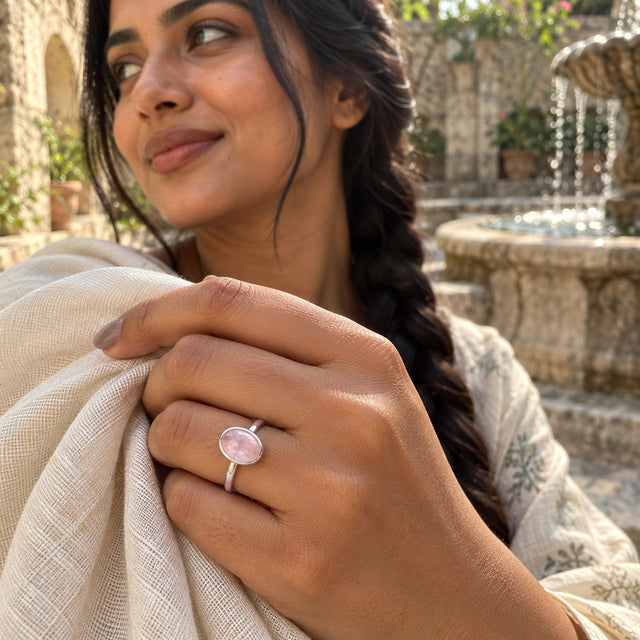 A silver rose quartz ring is shown on a hand holding ivory fabric beside a stone courtyard fountain.