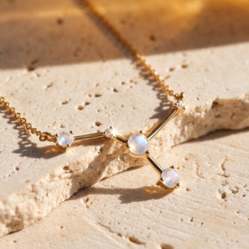 A yellow gold moonstone necklace is arranged diagonally on a pale stone surface in warm natural light.
